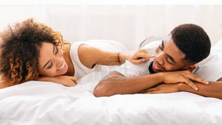Foto de Romance familiar. Atractiva mujer negra coqueteando con su marido, acostada en la cama, mirándose, sonriendo y hablando juntos - Imagen libre de derechos
