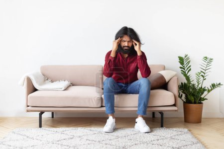 Indian man in a red shirt and jeans holding his head in pain while sitting on a sofa, with a plant beside him