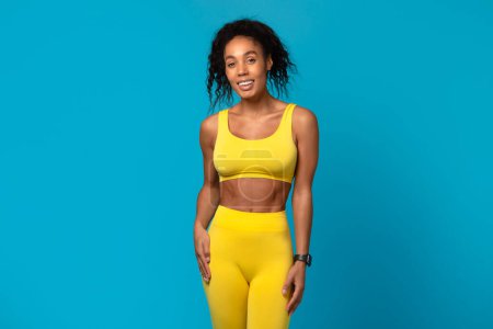 Téléchargez les photos : Jeune femme afro-américaine détendue et heureuse en tenue jaune sportive debout sur un fond bleu - en image libre de droit