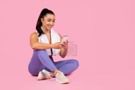 Téléchargez les photos : Femme sportive détendue naviguant sur son téléphone après une séance d'entraînement, entourée d'appareils de fitness - en image libre de droit