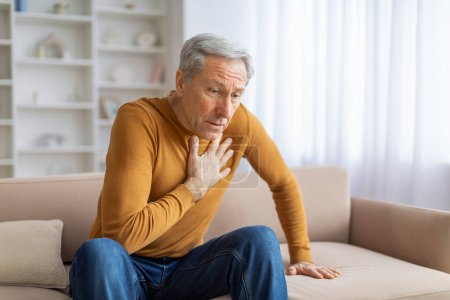 Hombre mayor parece en peligro mientras sostiene su pecho, posiblemente indicando un problema de salud, interior del hogar