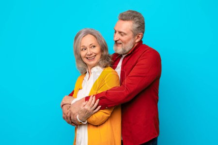Photo for Happy loving senior couple embracing with affection against blue backdrop in studio, showing love and bond between mature partners. Gray haired husband hugs his wife from back tenderly - Royalty Free Image