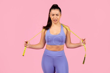 Photo for A young woman in sportswear appears distressed while measuring her waist with a measuring tape - Royalty Free Image