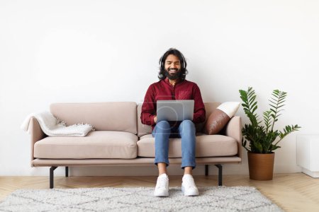 Glücklicher indischer Mann sitzt auf einem Sofa mit einem Laptop auf dem Schoß und strahlt Freude und Zufriedenheit in einem gut ausgestatteten Wohnzimmer aus