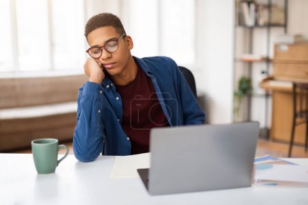 Müder schwarzer Student mit Brille und blauem Hemd döst an seinem Arbeitsplatz mit Laptop, Kaffeebecher und Notizbuch auf dem Schreibtisch