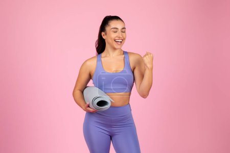 Photo for An energetic woman in workout gear holds a rolled-up yoga mat, smiling on a pink background, exuding happiness - Royalty Free Image