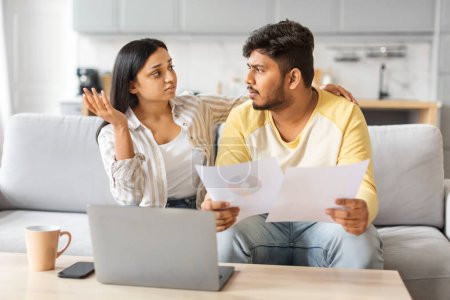 Photo for Indian man and woman are seated on a couch, focused on a piece of paper in their hands, stressed couple checking financial documents at home - Royalty Free Image