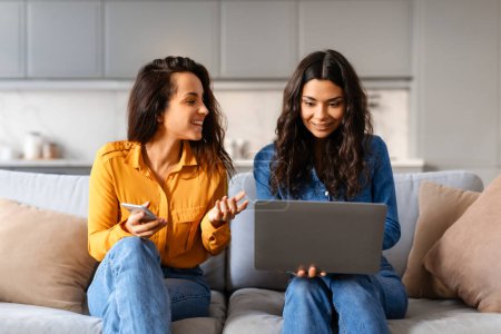 Dos mujeres sonrientes sentadas cómodamente en un sofá interactuando con una computadora portátil, posiblemente discutiendo trabajo o navegando por Internet