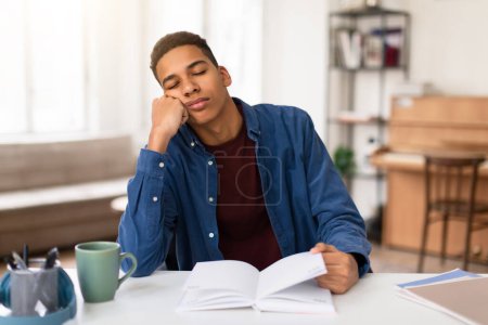 Photo for Fatigued black teen guy in blue shirt rests his head on his hand, dozing off while studying from textbook at white desk in bright room at home - Royalty Free Image