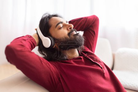 Téléchargez les photos : Un Indien confortable en chemise rouge faisant une pause et se relaxant sur un canapé dans un salon bien éclairé, écoutant de la musique - en image libre de droit