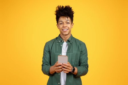 Téléchargez les photos : Jeune homme noir confiant dans une chemise verte tenant un téléphone portable, souriant avec un fond jaune - en image libre de droit