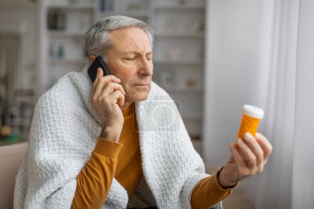 Concerned senior man looks at prescription bottle while using smartphone, consulting with doctor about medication