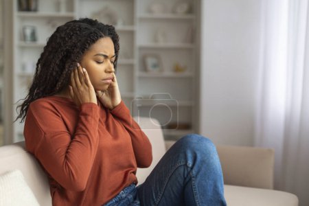 Photo for Adult black woman sitting on a couch holds her temples, african american female showing signs of discomfort, suffering acute headache, copy space - Royalty Free Image