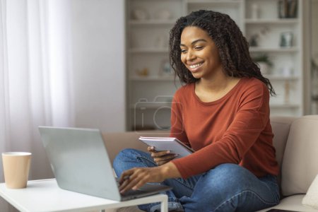 Téléchargez les photos : Femme noire joyeuse en vêtements décontractés à l'aide de son ordinateur portable tout en tenant un cahier, assis confortablement à la maison, afro-américaine femme travaillant ou étudier avec ordinateur à la maison - en image libre de droit
