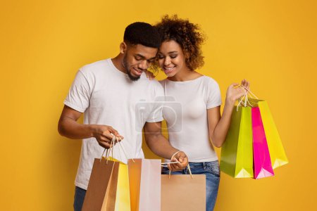 Foto de Sonriendo joven pareja afroamericana mirando en bolsas de compras con alegría contra un fondo amarillo vibrante, lo que sugiere una exitosa juerga de compras - Imagen libre de derechos
