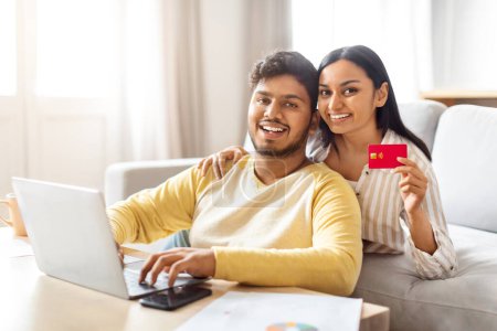 Téléchargez les photos : Indien homme et femme assis sur un canapé, tenant une carte de crédit rouge dans leurs mains et en utilisant un ordinateur portable, heureux couple shopping en ligne ensemble - en image libre de droit
