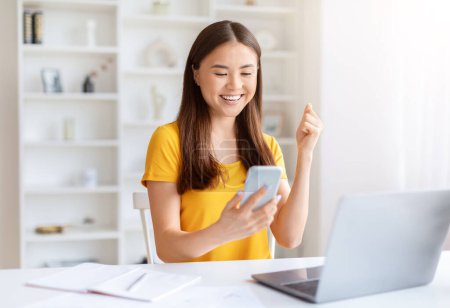 Photo for Ecstatic young asian woman celebrating good news on her smartphone at desk in front of laptop at home office - Royalty Free Image