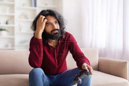 Téléchargez les photos : Homme indien avec les cheveux longs et la barbe assis sur le canapé montrant la déception tout en jouant, intérieur de la maison - en image libre de droit