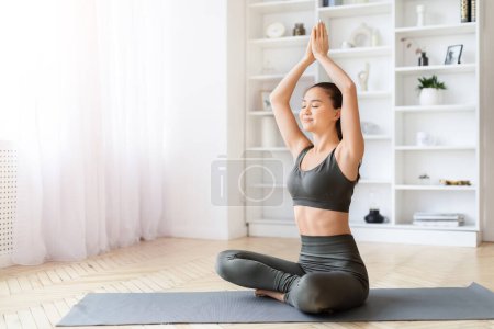 Smiling young asian woman enjoys yoga with hands in prayer pose in a cozy indoor setting, copy space