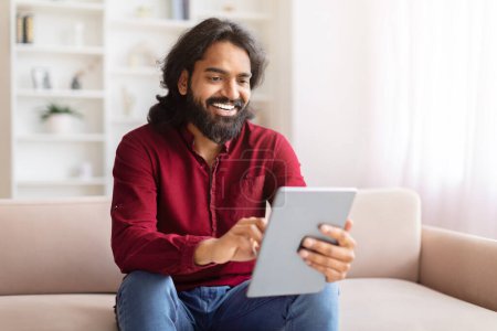Konzentrierter indischer Mann bequem mit einem digitalen Tablet, während er zu Hause auf einem Sofa sitzt und im Internet surft