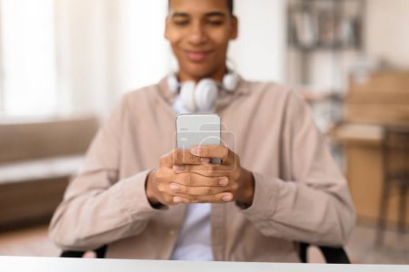 Photo for Cheerful black teenager guy with headphones around his neck smiles as he holds and looks at smartphone, engaging with technology at desk - Royalty Free Image
