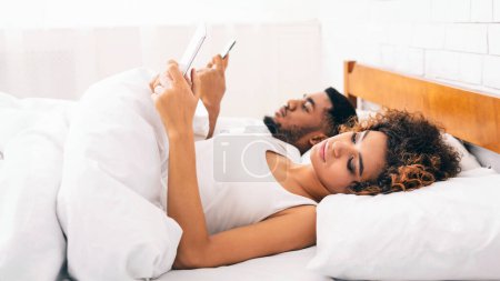 Photo for Workaholics family. Young black millennial couple with mobile phones sending emails, working from bed, empty space - Royalty Free Image