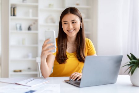 Mujer asiática feliz captura de una selfie con su teléfono inteligente por su ordenador portátil en el interior de la oficina en casa, espacio de copia