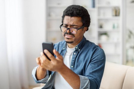 Téléchargez les photos : Homme noir plisse sur son téléphone avec une expression perplexe, peut-être dépanner un problème tout en étant assis sur le canapé dans le salon - en image libre de droit