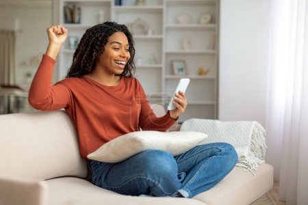 Foto de Una mujer negra sonriente sentada en un sofá levanta el puño triunfante mientras sostiene un teléfono móvil, una mujer afroamericana celebrando el éxito con un teléfono inteligente en casa - Imagen libre de derechos