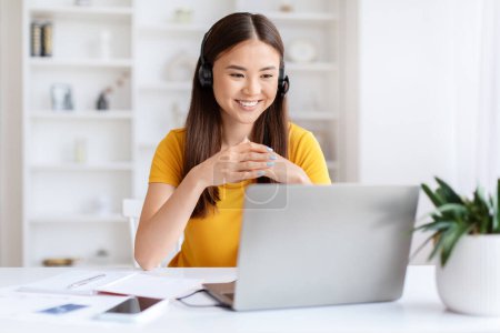 Foto de Mujer joven asiática con una sonrisa amigable usando auriculares y hablando con alguien a través de su computadora portátil - Imagen libre de derechos