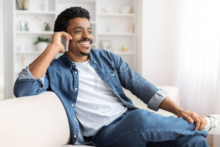 Foto de Un hombre negro está sentado en un sofá teniendo una conversación en su teléfono celular, sonriendo macho hablando en el teléfono móvil en casa - Imagen libre de derechos