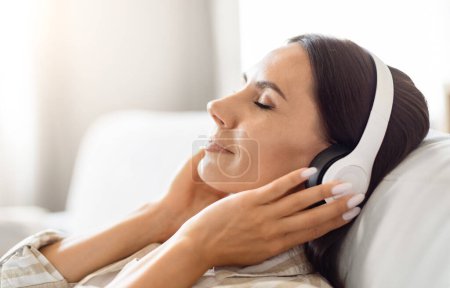 Téléchargez les photos : Une femme épuisée reposant sur un canapé, écouteurs allumés, dépeignant la relaxation avec une touche de technologie - en image libre de droit