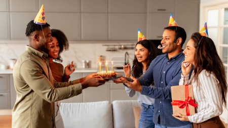 Multiracial friends holding a birthday cake with lit candles present it to a smiling woman during a cheerful celebration