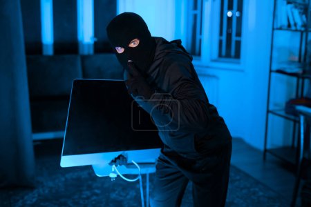 Voleur masqué portant une télévision ou un écran de bureau dans un appartement sombre, montrant un geste calme, portant des gants
