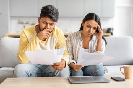 Photo for Stressed indian man and woman are seated on a couch, focusing on paperwork laid out in front of them. - Royalty Free Image