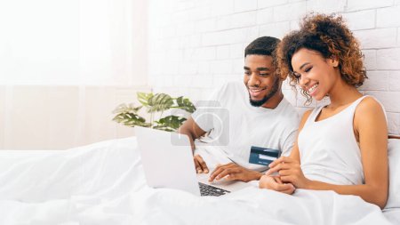 Téléchargez les photos : Afro-américain heureux couple utilise occasionnellement un ordinateur portable ensemble tout en étant assis dans le lit avec une carte de crédit à la main - en image libre de droit