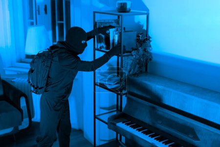 Photo for In a blue-lit room, a thief rummages through a shelving unit for valuables, with a piano in the foreground - Royalty Free Image