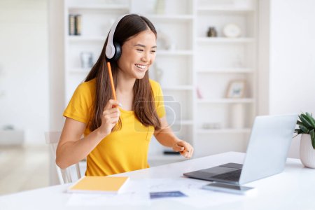 Téléchargez les photos : Une jeune femme asiatique joyeuse porte un casque tout en s'engageant avec un ordinateur portable dans un bureau à domicile lumineux - en image libre de droit