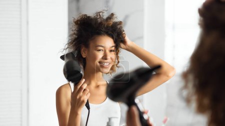 Foto de Secado por soplado. Sonriente mujer afroamericana secando el cabello con ventilador mirando en el espejo en el baño. Enfoque selectivo - Imagen libre de derechos