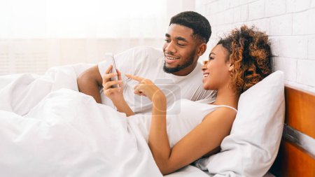 Foto de Pareja afroamericana acostada en la cama luciendo feliz y entretenida mientras comparte contenido en un teléfono móvil - Imagen libre de derechos