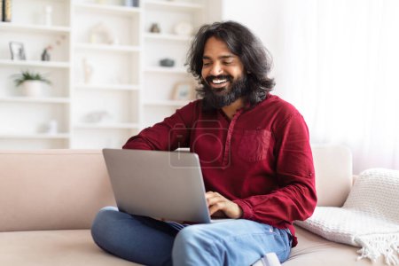 Indischer Mann sitzt auf einem pfirsichfarbenen Sofa und genießt seine Zeit bei der Arbeit oder unterhält sich mit einem Laptop in einem schön dekorierten Raum