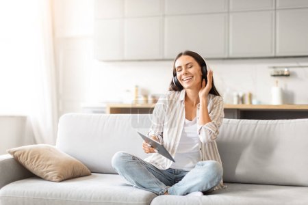 Eine entspannte Frau sitzt gemütlich auf einem Sofa und lächelt, während sie mit Tablet und Kopfhörer Musik hört.