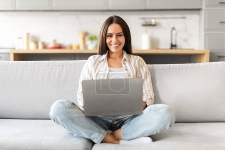 Mit überkreuzten Beinen auf der Couch lächelt eine Frau, als sie ihren Laptop in einem entspannten häuslichen Umfeld benutzt