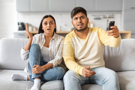 Photo for Indian couple seated on a couch displays shock and tension while watching television together, young couple emotionally reacting to content - Royalty Free Image