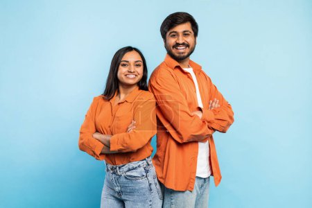 Téléchargez les photos : Deux jeunes hommes et femmes indiens en chemises orange assorties posant les bras croisés, souriant sur fond bleu - en image libre de droit