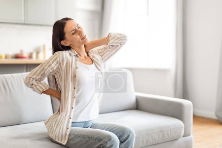 Téléchargez les photos : Femme brune assise sur le canapé à la maison, se frottant le cou et le bas du dos, sensation de crampes musculaires après la sieste, espace de copie - en image libre de droit