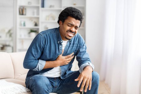 Photo for A concerned black male clutching his chest as he feels a sharp pain while sitting at home, feeling unwell - Royalty Free Image
