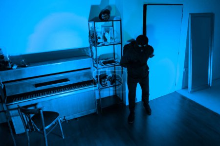 Photo for Masked burglar with a backpack inspects a dark room environment, planning their next move carefully - Royalty Free Image