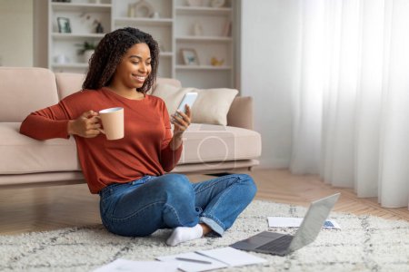 Photo for Smiling black woman sitting cross-legged on the floor with a cup of coffee and smartphone, african american female surrounded by documents and a laptop. - Royalty Free Image
