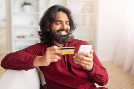 Foto de Un hombre indio encantado realiza de forma segura una compra en línea con su tarjeta de crédito y teléfono inteligente, retratando el comercio electrónico y la satisfacción - Imagen libre de derechos